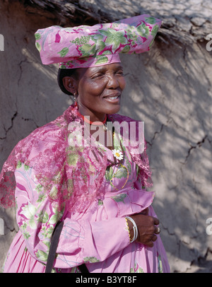 La Namibie, Damaraland, Palmwag. Une Femme Herero se tient dans l'ombre à l'extérieur de sa maison. Les origines de sa robe élaborée et unique style hat remonte au 19ème siècle les missionnaires allemands qui se sont opposés à ce qu'ils considéraient comme une forme de modestie parmi la tribu de robe. Ils ont insisté pour que les femmes Herero peu pratique d'adopter leur propre style de robes de l'époque victorienne. Au fil des ans, robes Herero colorés ont évolué aussi énorme crinolines porté sur cinq jupons. Banque D'Images