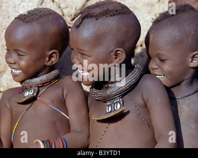 La Namibie, Kaokoland, Epupa. Trois heureux enfants Himba - deux filles et un garçon - regarder une danse. Les ornements des filles : colliers de perles blanches, appelé ombwari, une tradition de toutes les personnes et Himba perles en métal soutenu sur le cuir, qui sont ornés de cauris issue de l'Afrique de la côte Atlantique.Les Himbas Herero Bantu francophone sont des nomades qui vivent dans les conditions difficiles, sec mais très beau paysage de la nord-ouest de la Namibie. Banque D'Images