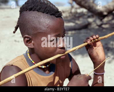 La Namibie, Kaokoland, Epupa. Un jeune Himba, son style de cheveux dans une longue natte appelée ondatu, joue un arc - bouche d'un instrument de musique privilégié par les jeunes garçons. Il est fabriqué à partir du palmier Makalani.Les Himbas Herero Bantu francophone sont des nomades qui vivent dans les conditions difficiles, sec mais très beau paysage de la nord-ouest de la Namibie. Banque D'Images