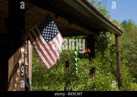 Un drapeau américain en lambeaux s'agite dans la brise sur le porche d'une vieille maison de bois, avec des ornements du vent derrière. Banque D'Images