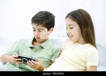 Frère et sœur siégeant ensemble, boy playing handheld video game Banque D'Images