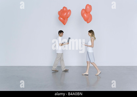 Jeune homme et femme marche vers l'autre, à la fois garder leurs ordinateurs portables et ballons coeur Banque D'Images