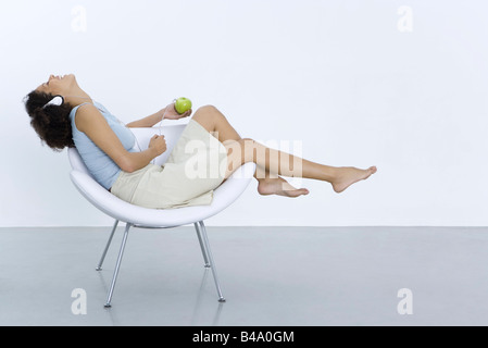 Woman reclining in chair, l'écoute au casque connecté à apple, side view Banque D'Images