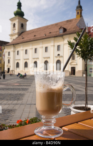 Café Latte Macchiato avec de la paille dans le verre sur la table à café de la rue dans la ville historique de Piata Mare, Sibiu, Transylvanie, Roumanie, Europe. Banque D'Images