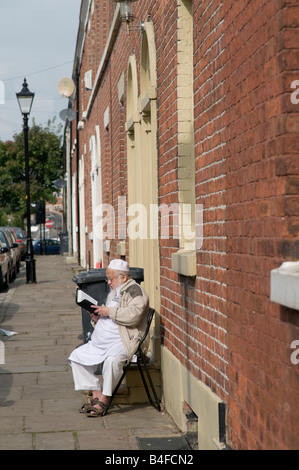 Personnes âgées homme musulman lecture livre religieux assis sur une chaise à l'extérieur de sa maison mitoyenne à Preston Lancashire England UK Banque D'Images