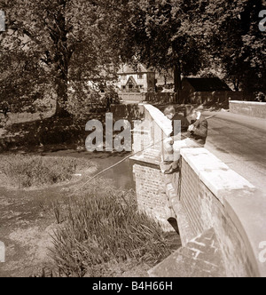 Deux jeunes garçons assis sur le côté d'un pont comme ils essaient de pêcher dans leur rivière locale à l'aide d'un bâton long circa 1950 Banque D'Images