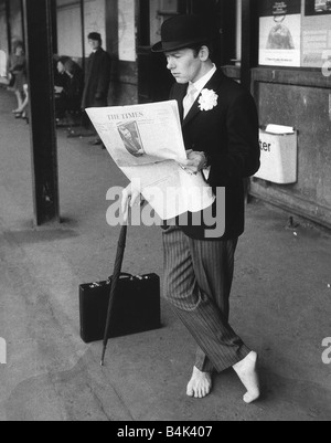 Une ville gent habillé de rayures broche chapeau melon et s'appuyant sur un lit son brolley copie du journal The Times sur une gare ferroviaire de la plate-forme de l'homme n'est pas de porter des chaussettes, et des chaussures Banque D'Images