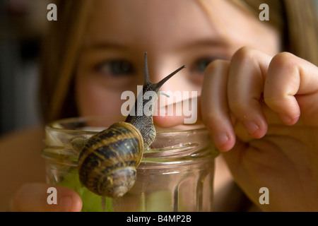 Une jeune fille de race blanche regarde un jardin commun (escargots Helix aspersa) grimper sur son doigt, qui repose sur le dessus d'un bocal de verre. Banque D'Images