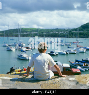 Un garçon assis sur le quai de pierre à la recherche de bateaux dans le port New Quay West Wales Royaume-uni CEREDIGION KATHY DEWITT Banque D'Images