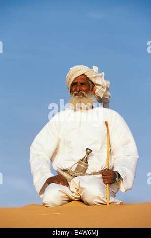 Oman, Sharqiyah, Wahiba Sands. Un Bédouin l'homme s'agenouille au sommet d'une dune de sable dans le désert. Il porte la robe traditionnelle omanaise Banque D'Images