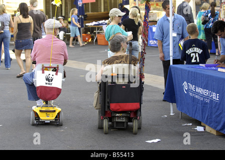 Un couple de personnes âgées bénéficie d'un voyage à un événement public à leur mobilité scooter et fauteuil roulant motorisé. Banque D'Images