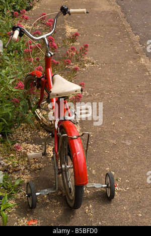 Une ancienne bicyclette d'enfant rouge avec des roues stabilisatrices sur le trottoir d'une rue tranquille, Banque D'Images