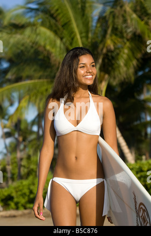 Une jolie jeune femme dans un bikini blanc est titulaire d'un surf à Lahaina, Maui, Hawaï. Banque D'Images