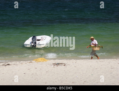 Un vendeur de fruits tunisien promenades le long d'une plage de sable fin à la mer Méditerranée, un bateau blanc se trouve ancré dans l'eau. Banque D'Images