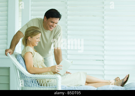 Woman relaxing in chair, livre de lecture, l'homme penché sur son épaule et souriant Banque D'Images