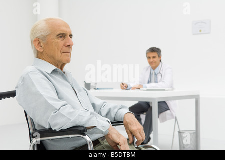 Senior patient assis en fauteuil roulant, un médecin travaillant à 24 en arrière-plan Banque D'Images