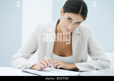 Young businesswoman en utilisant un stylo pour saisir des chiffres sur calculatrice, looking at camera Banque D'Images