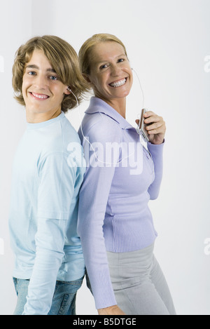 Mère et fils adolescents listening to MP3 player ensemble, debout dos à dos, smiling at camera Banque D'Images