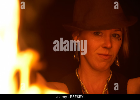 Femme firepoise fire dance performance artist wearing hat holding fire fans regardant à travers les flammes dans la nuit Banque D'Images