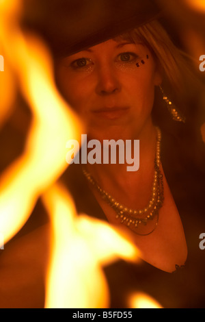 Femme firepoise fire dance performance artist wearing hat holding fire fans regardant à travers les flammes dans la nuit Banque D'Images