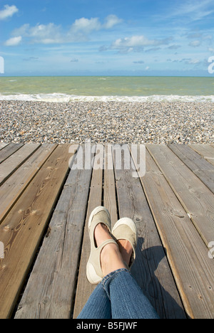 Vue de deux jambes et les pieds reposant sur un conseil marche à côté d'une plage de galets et de l'océan Banque D'Images