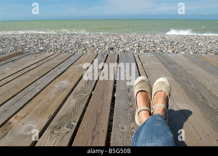 Vue de deux jambes et les pieds reposant sur un conseil marche à côté d'une plage de galets et de l'océan Banque D'Images