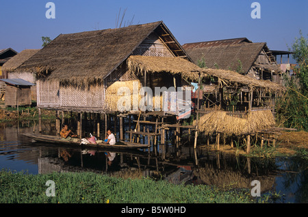 Les femmes et les enfants birmans dans un canoë glissent au-delà de leur hutte de chaume ou de la maison de stilt soulevée sur les pilotis Inle Lake Birmanie ou Myanmar Banque D'Images