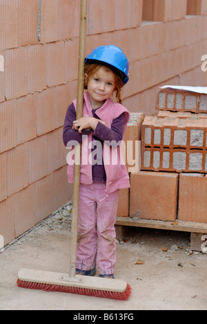 Little girl holding a broom et portant un casque bleu à une maison construction site Banque D'Images