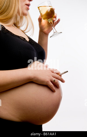 A 8 mois et demi de femme enceinte fume une cigarette et de boire un verre de vin blanc en studio contre fond uni, UK Banque D'Images