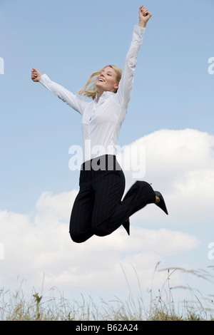 Jeune femme blonde portant des vêtements d'affaires de sauter devant un ciel bleu, les bras tendus Banque D'Images