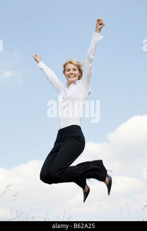 Jeune femme blonde portant des vêtements d'affaires de sauter devant un ciel bleu, les bras tendus Banque D'Images