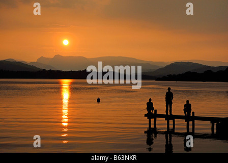 Trois jeunes hommes, deux assis, l'un debout, sur une jetée regardant le coucher de soleil sur le lac Windermere, Lake District Banque D'Images