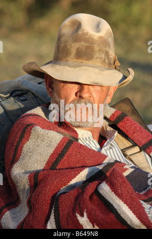 Un vieux cowboy minuterie appuyé contre son sac à côté de la selle et chuck wagon au coucher du soleil Banque D'Images