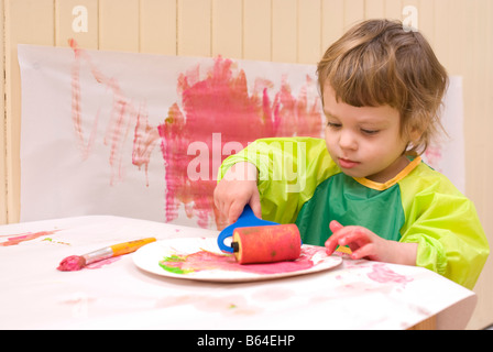 Une fillette de deux ans dans un plastique smock peinture dans un jeu avec un pinceau et rouleau Banque D'Images