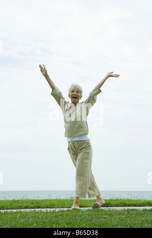 Senior woman standing outdoors in seaside park avec bras levés, sourire heureux Banque D'Images