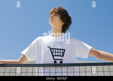 Teenage boy wearing tee-shirt imprimé avec panier, debout à balustrade, looking up Banque D'Images