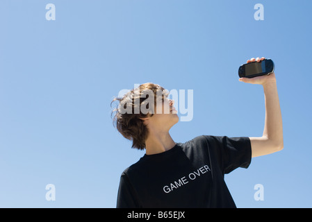 Teen boy holding up jeu vidéo de poche, portant des tee-shirt imprimé avec les mots 'game over' Banque D'Images