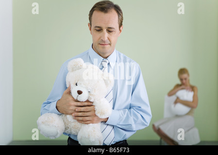 Nouveau teddy bear père, regardant vers le bas, l'épouse holding baby in background Banque D'Images