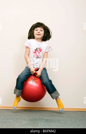 Petite fille, 6 ans, de sauter sur une boule rouge, brune, de l'intérieur Banque D'Images