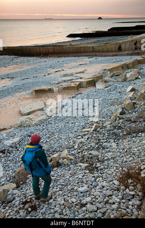 Femme randonneur sur plage au coucher du soleil avec un mur de défense de la mer et des brise-lames Aberthaw Wales UK Banque D'Images