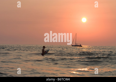 Garçon natation en eau peu profonde au coucher du soleil avec voilier en arrière-plan Banque D'Images