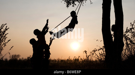 Silhouette d'enfants indiens jouant sur une balançoire fait maison à la campagne au coucher du soleil. L'Andhra Pradesh, Inde Banque D'Images