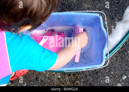 Fillette de deux ans s'amusant à jouer avec de l'eau savonneuse tout en aidant à se laver la voiture familiale Banque D'Images