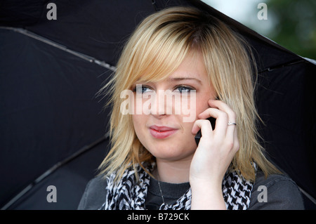 Fille de 18 ans blonde holding umbrella à sourire de l'écoute de téléphone mobile Banque D'Images