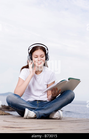 Jeune adolescent assis sur le pont, le port de sortie casque holding book smiling portrait Banque D'Images