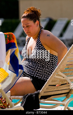 Palm Beach Shores , grande dame d'âge moyen corpulent avec tatouage en maillot de bain sur une chaise longue au soleil au bord de piscine Banque D'Images