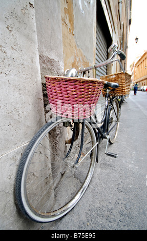 Vélo avec panier Rome Italie | ROME, Italie — Un vélo noir équipé d'un panier avant en osier rose et d'un panier arrière plus petit s'appuie contre un mur en stuc altéré sur une rue romaine. Les paniers tissés restent un accessoire pratique commun sur les vélos de ville, prisé pour transporter des épiceries et de petits colis dans des voies étroites et restreintes pour les voitures. Une deuxième bicyclette en panier se trouve plus loin le long de la même paroi. Banque D'Images