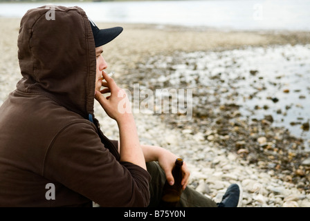 Lonely Boy est situé sur rive et boit de la bière d'une bouteille et fume une cigarette Banque D'Images