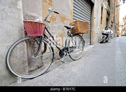 Vélo avec panier Rome Italie | ROME, Italie — Un vélo avec des paniers en osier rose et naturel assortis repose contre un mur en stuc qui s'effrite dans une rue calme romaine, un volet roulant fermé et une moto garée visible derrière. La scène reflète l'utilisation quotidienne des pédales pour les courses dans les quartiers plus anciens et plus étroits de la ville, où l'accès aux voitures est souvent limité. Banque D'Images