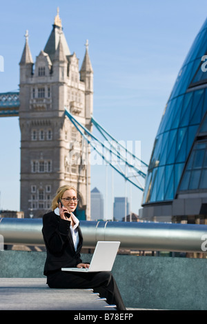 Une belle jeune femme executive en utilisant son ordinateur portable et téléphone mobile en face de Tower Bridge Canary Wharf et la ville de Londres Banque D'Images